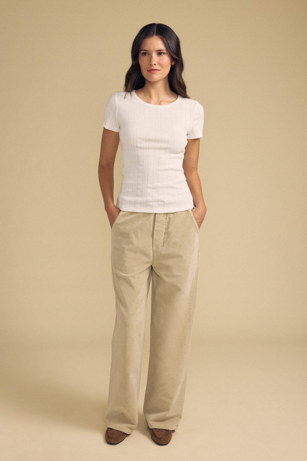 A woman stands with her hands in her pockets, wearing the LENA POINTELLE TEE by Velvet by Graham & Spencer and loose beige pants, set against a plain beige background.-41834091741377