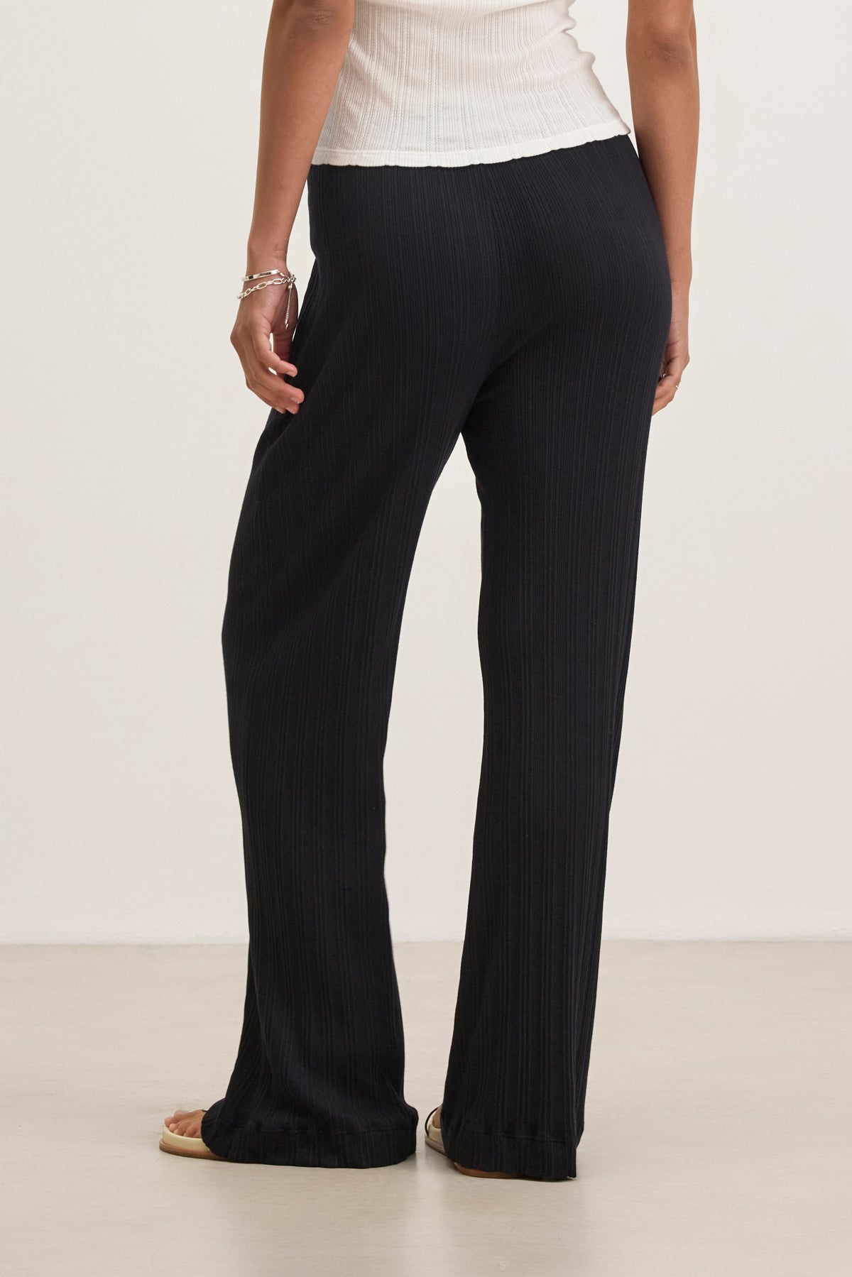 A person faces away from the camera, wearing a white top and ALEMI POINTELLE PANT by Velvet by Graham & Spencer, known for their flattering fit. They stand on a light-colored surface with one hand resting by their side.-38802035048641