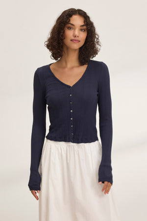 A person with curly hair wears the DREW POINTELLE CARDIGAN by Velvet by Graham & Spencer, a dark, fitted cotton-modal blend cardigan, paired with a white skirt against a plain background.