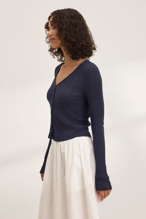 A woman with curly hair wears the Velvet by Graham & Spencer DREW POINTELLE CARDIGAN in navy, paired with a white skirt, standing in profile against a plain backdrop, showcasing an elegant layered look.