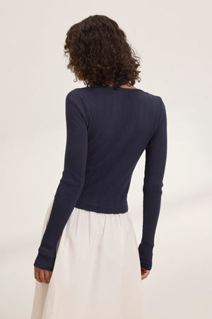 The individual elegantly showcases layering by wearing Velvet by Graham & Spencer's navy DREW POINTELLE CARDIGAN and a white skirt, standing with their back to the camera against a plain backdrop.