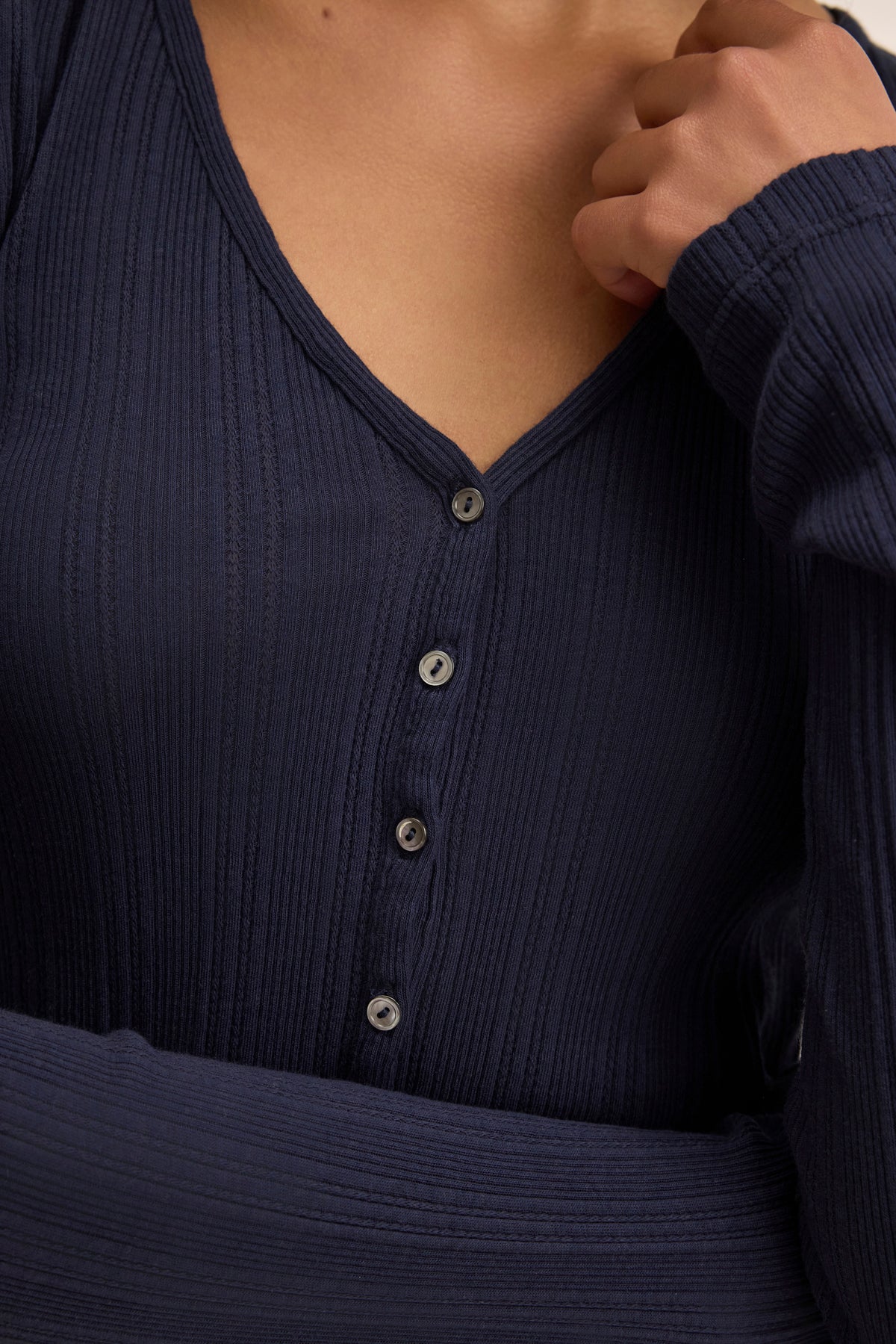 A person in a navy blue ribbed V-neck top made from a soft cotton-modal blend, featuring front buttons, holds the collar. Ideal for layering with your DREW POINTELLE CARDIGAN by Velvet by Graham & Spencer.-38802080694465