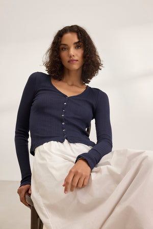 A woman with curly hair sits on a chair against a plain background, wearing the DREW POINTELLE CARDIGAN by Velvet by Graham & Spencer over a dark buttoned top and light skirt. This cozy ensemble, ideal for layering, is crafted from a cotton-modal blend.