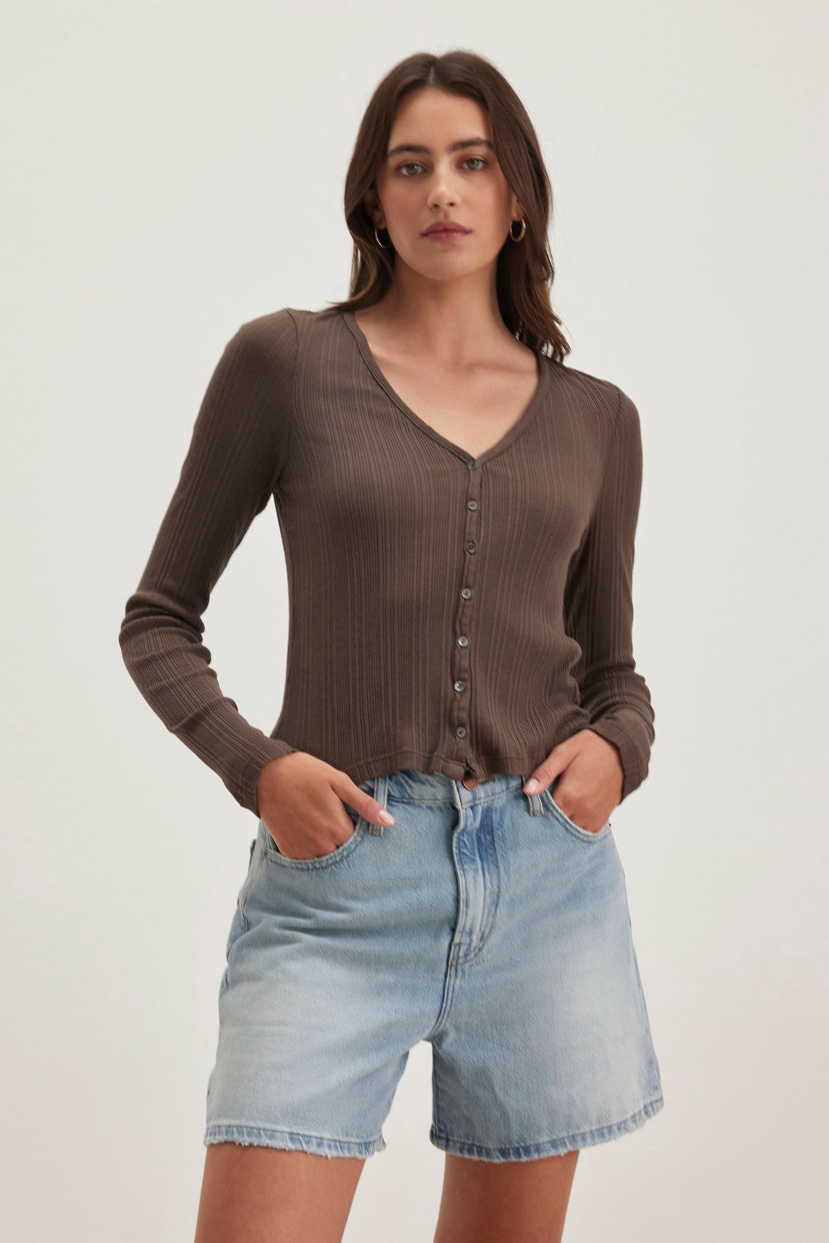 The person flaunts a DREW POINTELLE CARDIGAN by Velvet by Graham & Spencer, paired with light blue denim shorts, standing hands-in-pockets against a plain background to showcase casual layering effortlessly.