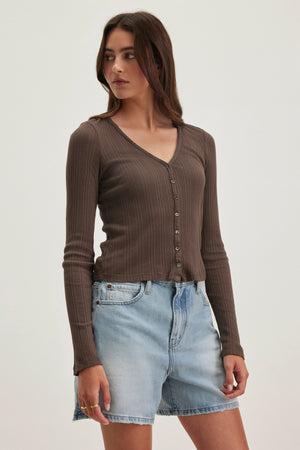 A woman models a ribbed, long-sleeve brown top with buttons and light blue denim shorts. Her side-gazing stance against a plain backdrop highlights the ensemble as ideal for layering with Velvet by Graham & Spencer's DREW POINTELLE CARDIGAN. Her long brown hair complements the casual look.