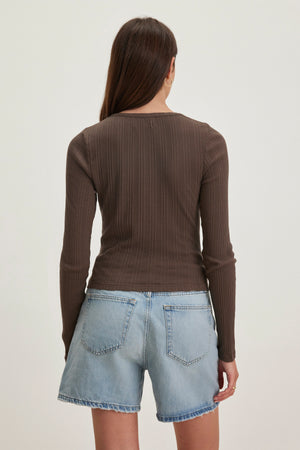 Rear view of a person with long brown hair wearing the Velvet by Graham & Spencer's DREW POINTELLE CARDIGAN in brown, perfect for layering, paired with blue denim shorts.
