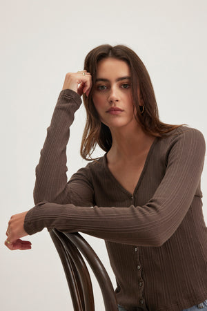 A woman with long brown hair in the Velvet by Graham & Spencer DREW POINTELLE CARDIGAN leans on a wooden chair against a neutral background, effortlessly showcasing the perfect layering look.