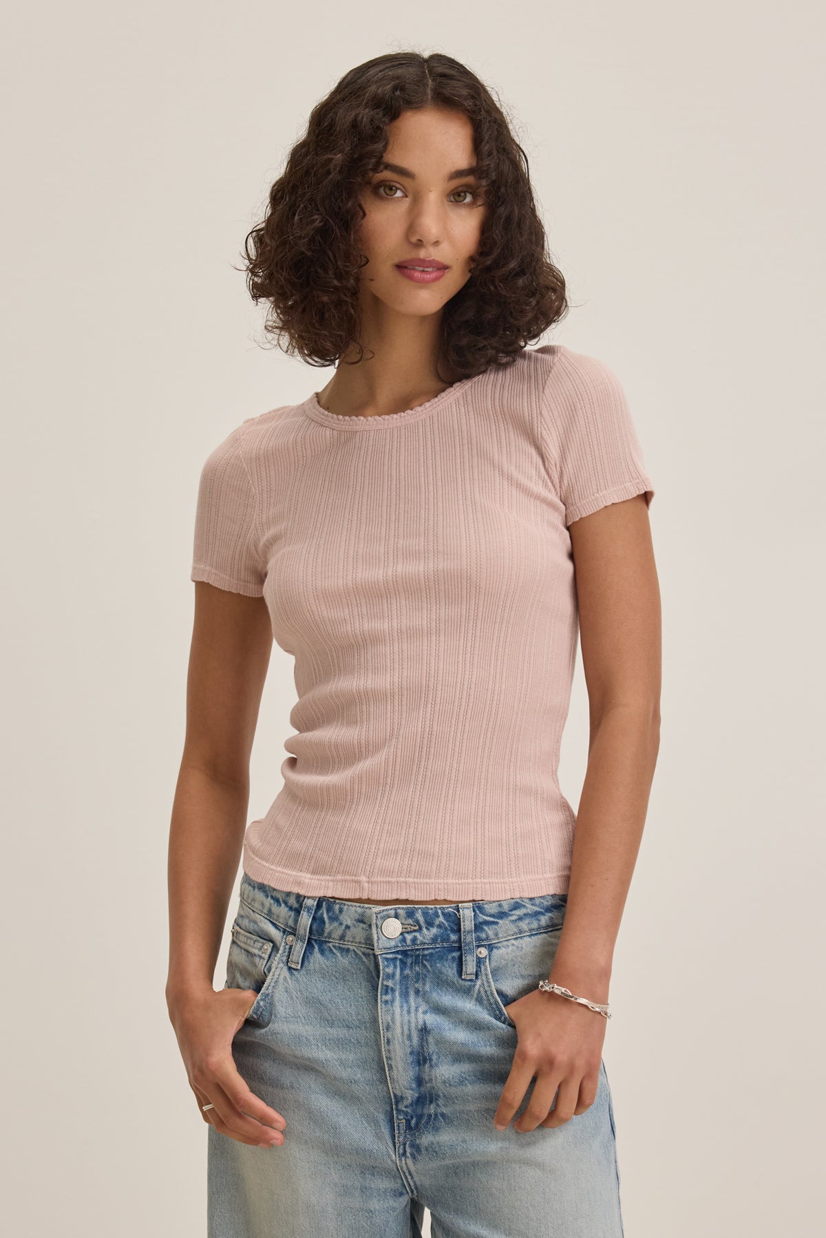 A person with curly hair is wearing a pink ENATI POINTELLE TEE by Velvet by Graham & Spencer and blue jeans, standing casually with hands in pockets against a plain background.-38820606869697