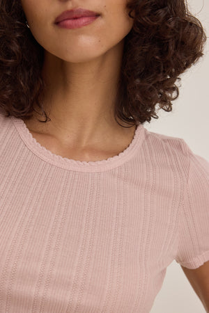 A person with shoulder-length curly hair is wearing the ENATI POINTELLE TEE by Velvet by Graham & Spencer, a light pink ribbed shirt with a scalloped neckline.