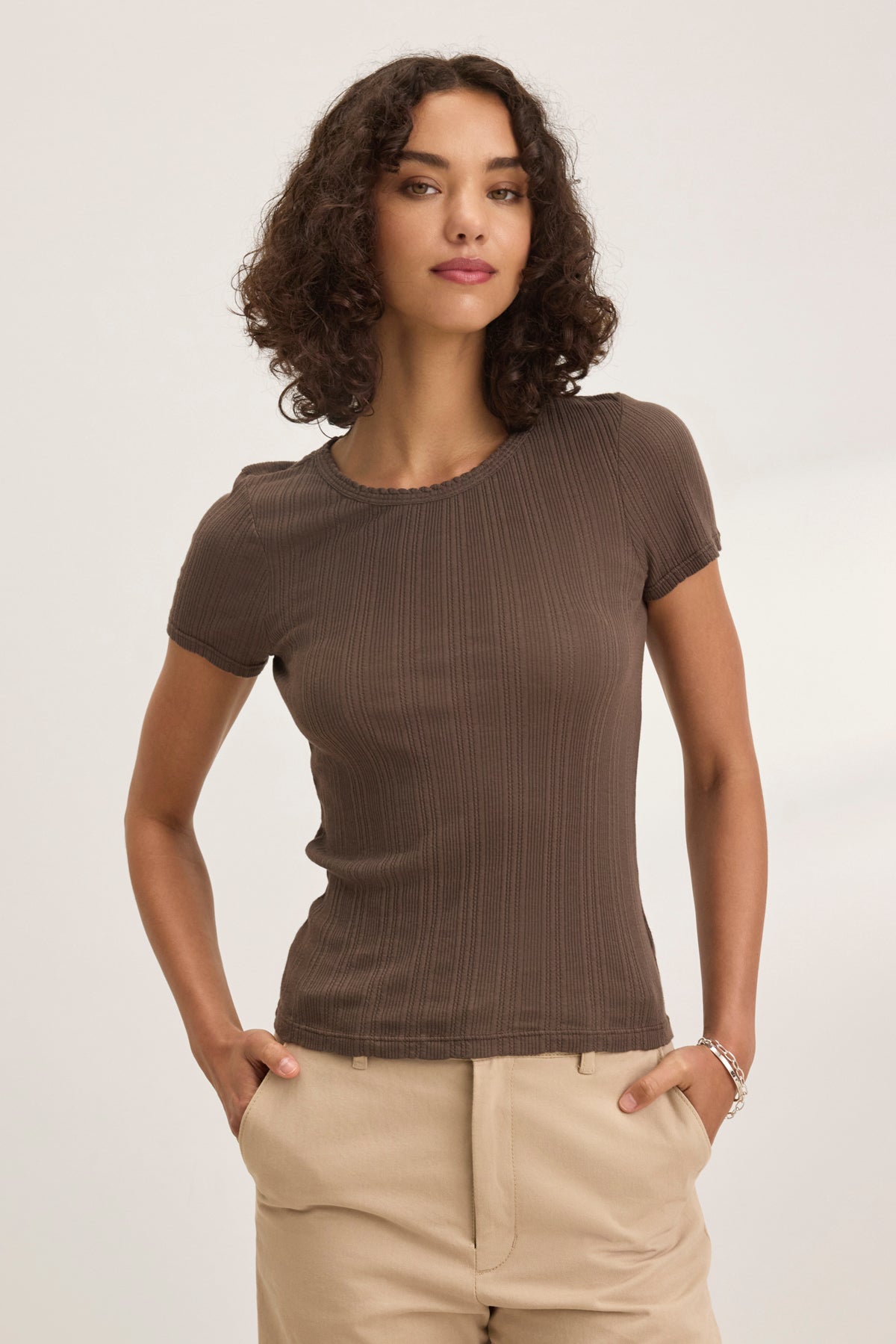 A woman with curly hair wears the ENATI POINTELLE TEE from Velvet by Graham & Spencer in brown, paired with beige pants, standing hands-in-pockets against a plain backdrop.-38820606673089