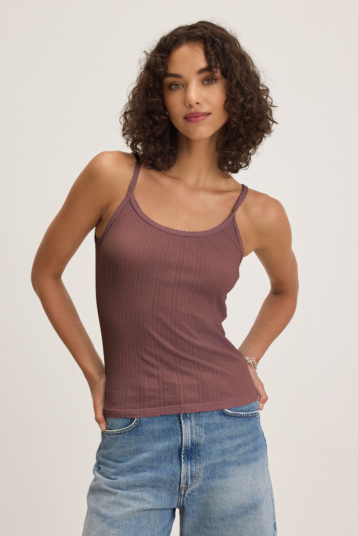 A woman with curly hair wears the INIKU POINTELLE TANK TOP by Velvet by Graham & Spencer in mauve with light blue jeans, standing against a plain background—a versatile wardrobe staple.