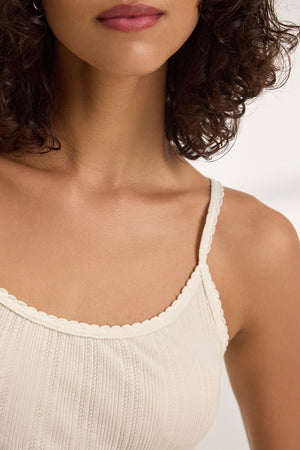 Close-up of a person with curly hair wearing the INIKU POINTELLE TANK TOP by Velvet by Graham & Spencer, showcasing a cream color with a ribbed texture and scalloped edges.
