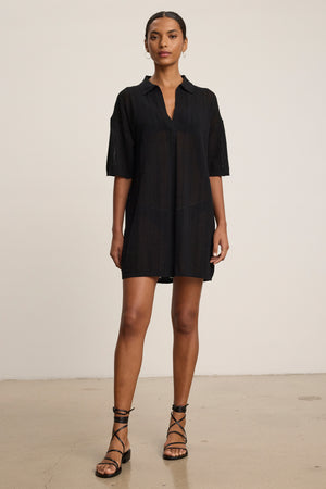 A woman stands indoors in the MILA POINTELLE LACE DRESS by Velvet by Graham & Spencer and black strappy sandals, posed against a plain light background—an effortless addition to any summer wardrobe.