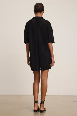 A person stands indoors facing away from the camera, wearing the MILA POINTELLE LACE DRESS by Velvet by Graham & Spencer with black sandals—a versatile addition to any summer wardrobe.
