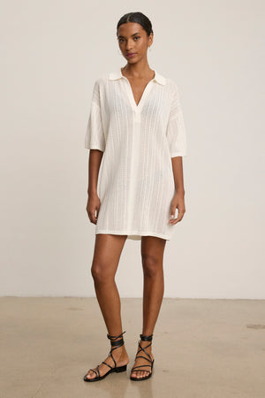 A woman stands indoors facing forward against a plain background, wearing the MILA POINTELLE LACE DRESS by Velvet by Graham & Spencer with a collar and white pointelle lace cotton fabric, paired with black strappy sandals.