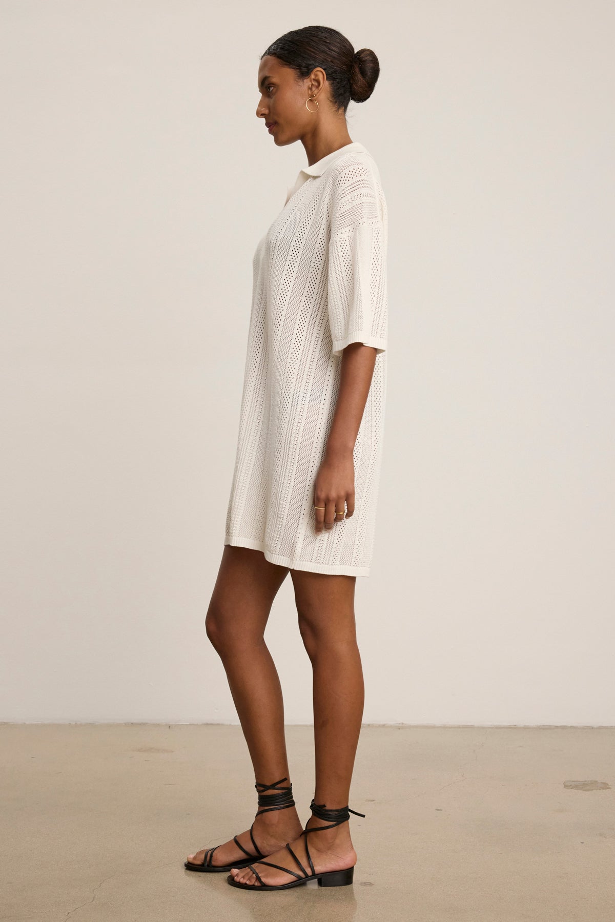 A woman stands in profile wearing the MILA POINTELLE LACE DRESS by Velvet by Graham & Spencer—a knee-length white cotton sweater polo dress with half sleeves—paired with black lace-up sandals, set against a plain background.