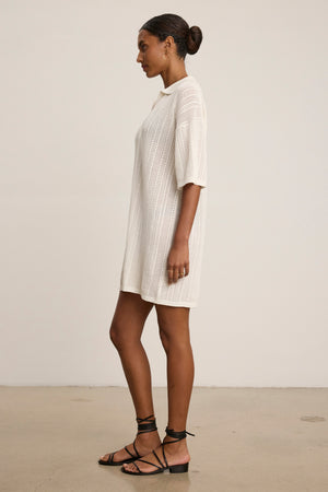 A woman stands in profile wearing the MILA POINTELLE LACE DRESS by Velvet by Graham & Spencer—a knee-length white cotton sweater polo dress with half sleeves—paired with black lace-up sandals, set against a plain background.