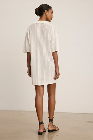 A woman, seen from behind, wears the MILA POINTELLE LACE DRESS by Velvet by Graham & Spencer—a cream short-sleeve sweater polo dress with lace cotton details—paired with black strappy sandals against a plain background.