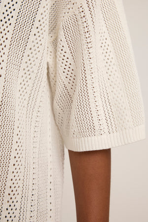 A close-up of a person's arm wearing the MILA POINTELLE LACE DRESS by Velvet by Graham & Spencer, featuring a cream-colored cotton knit with short sleeves and an openwork lace pattern.