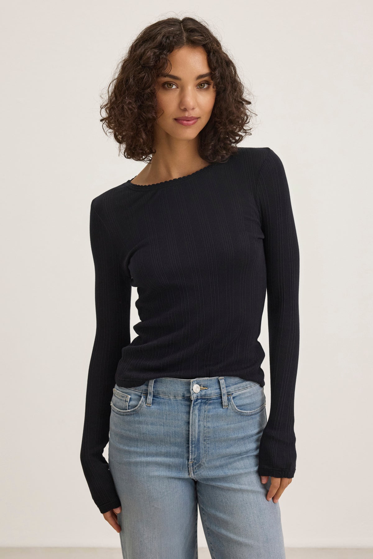 A curly-haired woman wearing a MAURI POINTELLE TEE by Velvet by Graham & Spencer in black and blue jeans stands against a plain background.-38820767662273