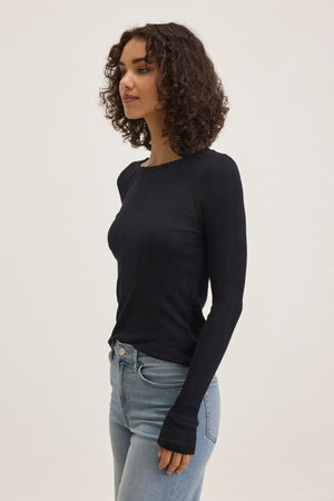A person with curly hair wears the MAURI POINTELLE TEE by Velvet by Graham & Spencer, a long-sleeved black top with ribbed pointelle accents, paired with blue jeans. They stand against a plain background, highlighting the subtle elegance of their outfit.