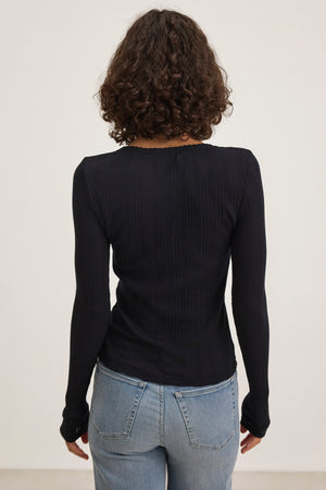 The rear view shows a person with curly hair wearing Velvet by Graham & Spencer's MAURI POINTELLE TEE, a black long-sleeve tee with ribbed pointelle accents, paired with blue jeans against a plain background.
