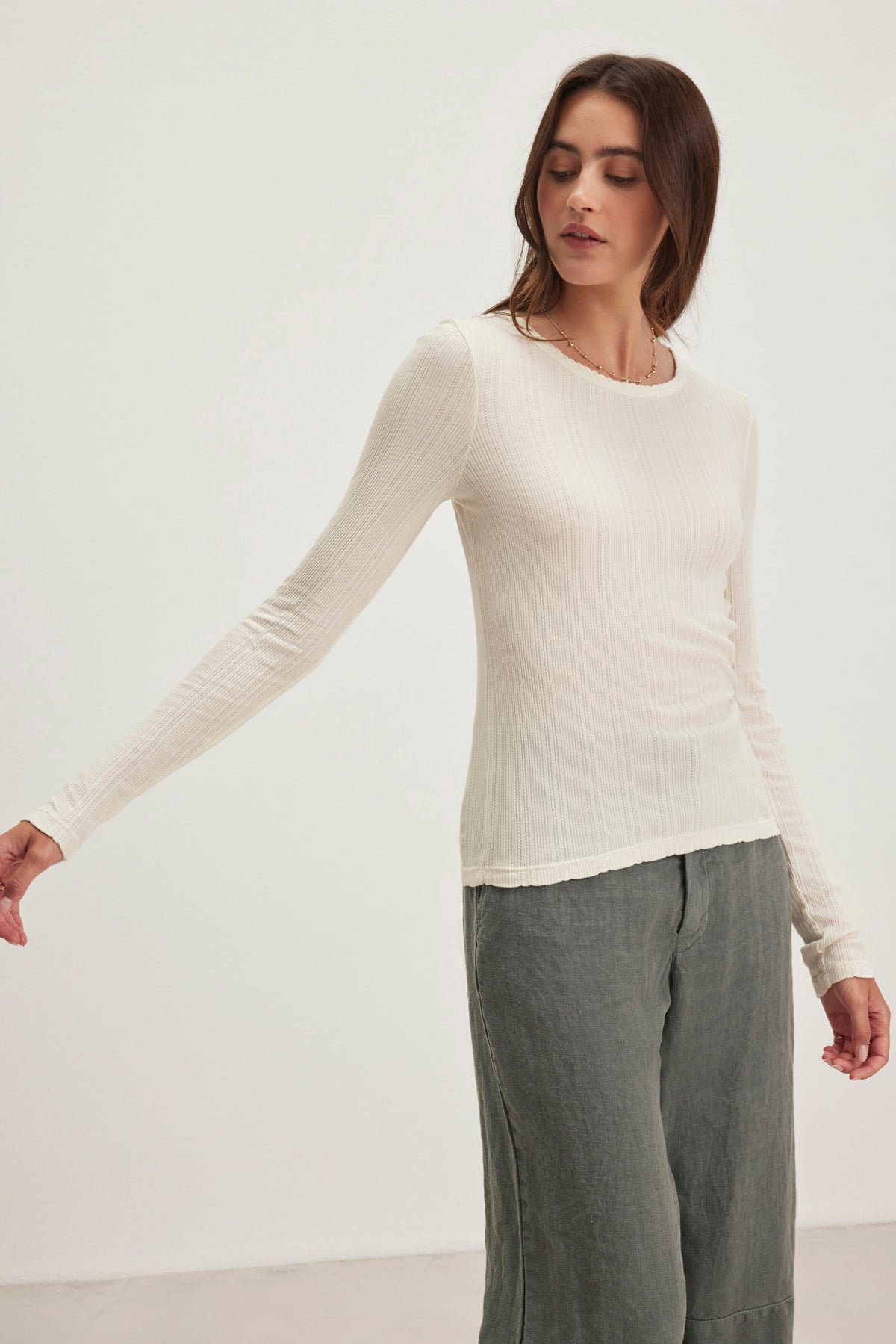 A person wearing the MAURI POINTELLE TEE by Velvet by Graham & Spencer, featuring a white long-sleeve with ribbed pointelle accents, and gray pants stands against a plain background with one arm gently extended while looking downward.-38820768121025
