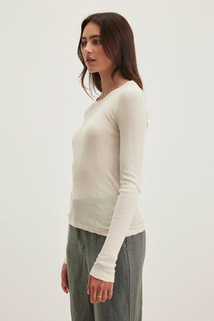 A person stands in profile against a plain background, wearing the Velvet by Graham & Spencer MAURI POINTELLE TEE, a white ribbed long-sleeve shirt with pointelle accents, paired with gray pants.