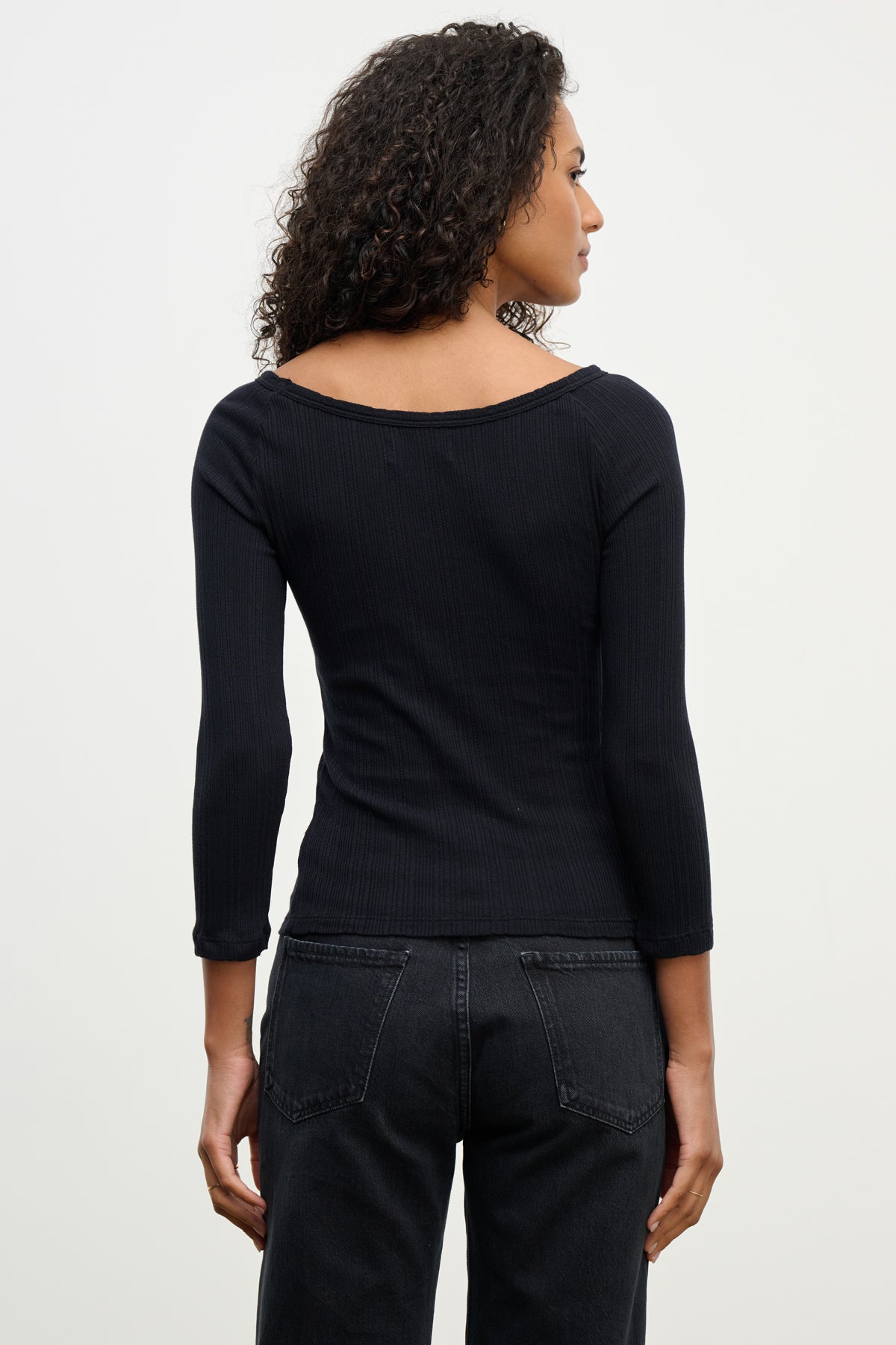 A person with curly hair is seen from behind, wearing the Velvet by Graham & Spencer NIMA POINTELLE TEE in black with black jeans, standing against a plain light background.-39742363173057