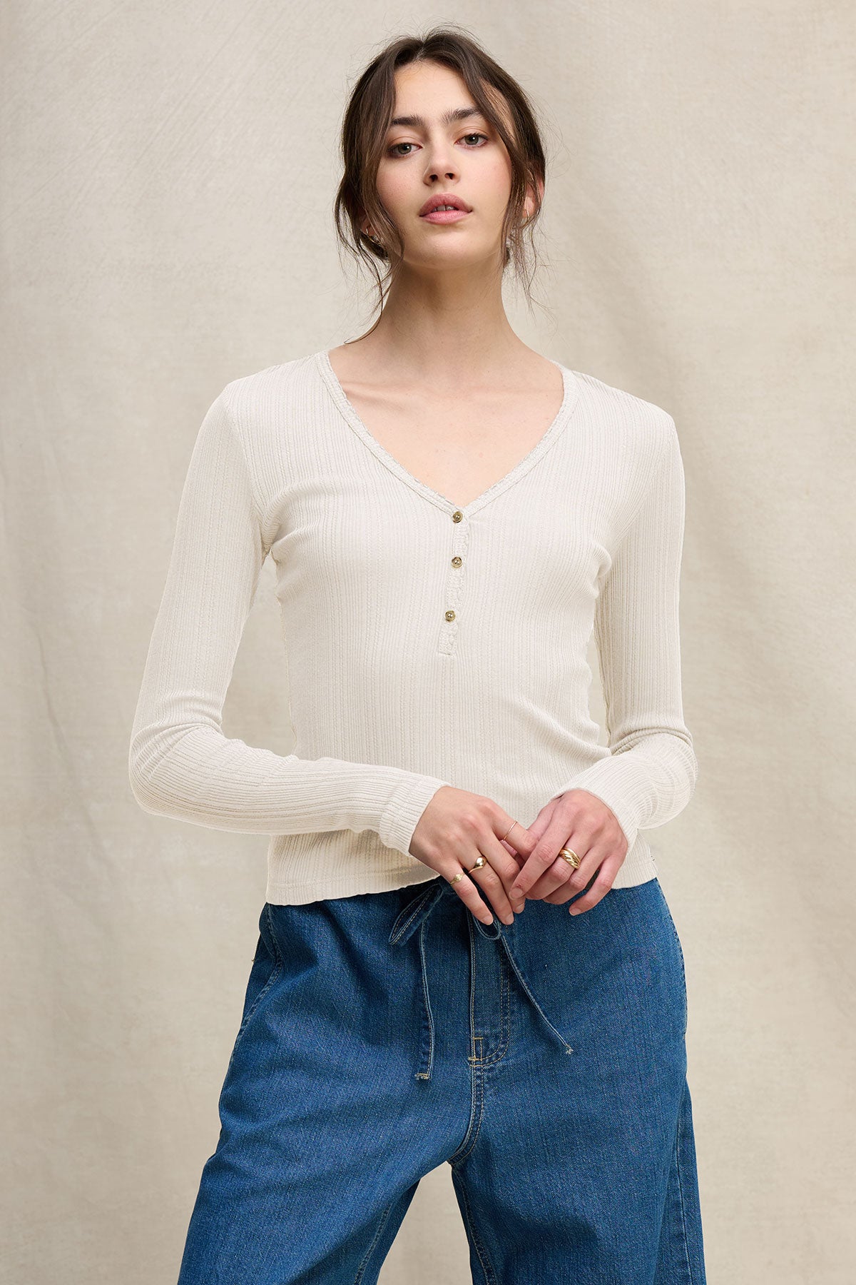 A woman wears the WINONA POINTELLE HENLEY by Velvet by Graham & Spencer, a cream long-sleeve V-neck top with a scalloped neckline, paired with blue drawstring jeans, standing against a neutral background.