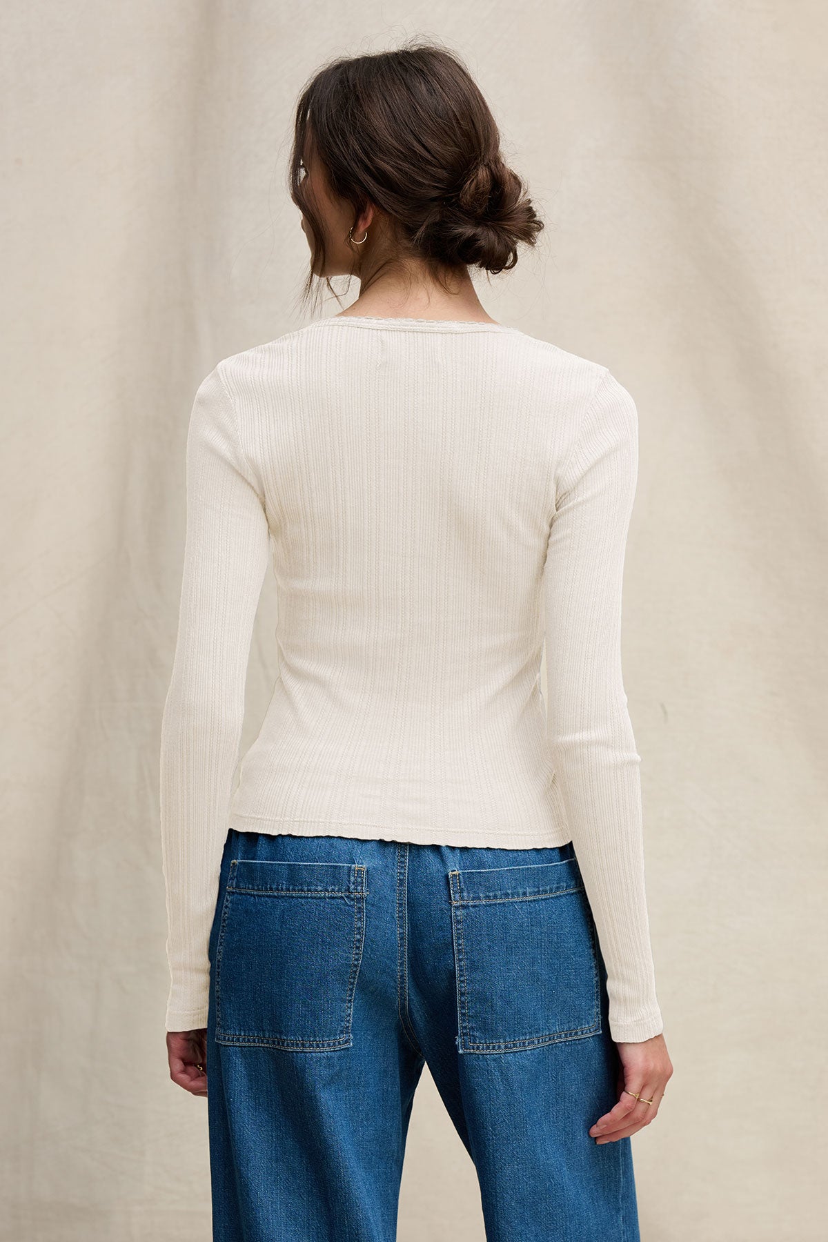 A woman stands facing away from the camera, wearing the WINONA POINTELLE HENLEY by Velvet by Graham & Spencer and blue denim jeans with large back pockets.
