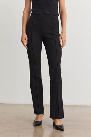 The individual is dressed in the ALBEE PONTE PANT from Velvet by Graham & Spencer, featuring a high-waisted and boot-cut silhouette, paired with a sleeveless top and black pointed shoes on a plain floor background.