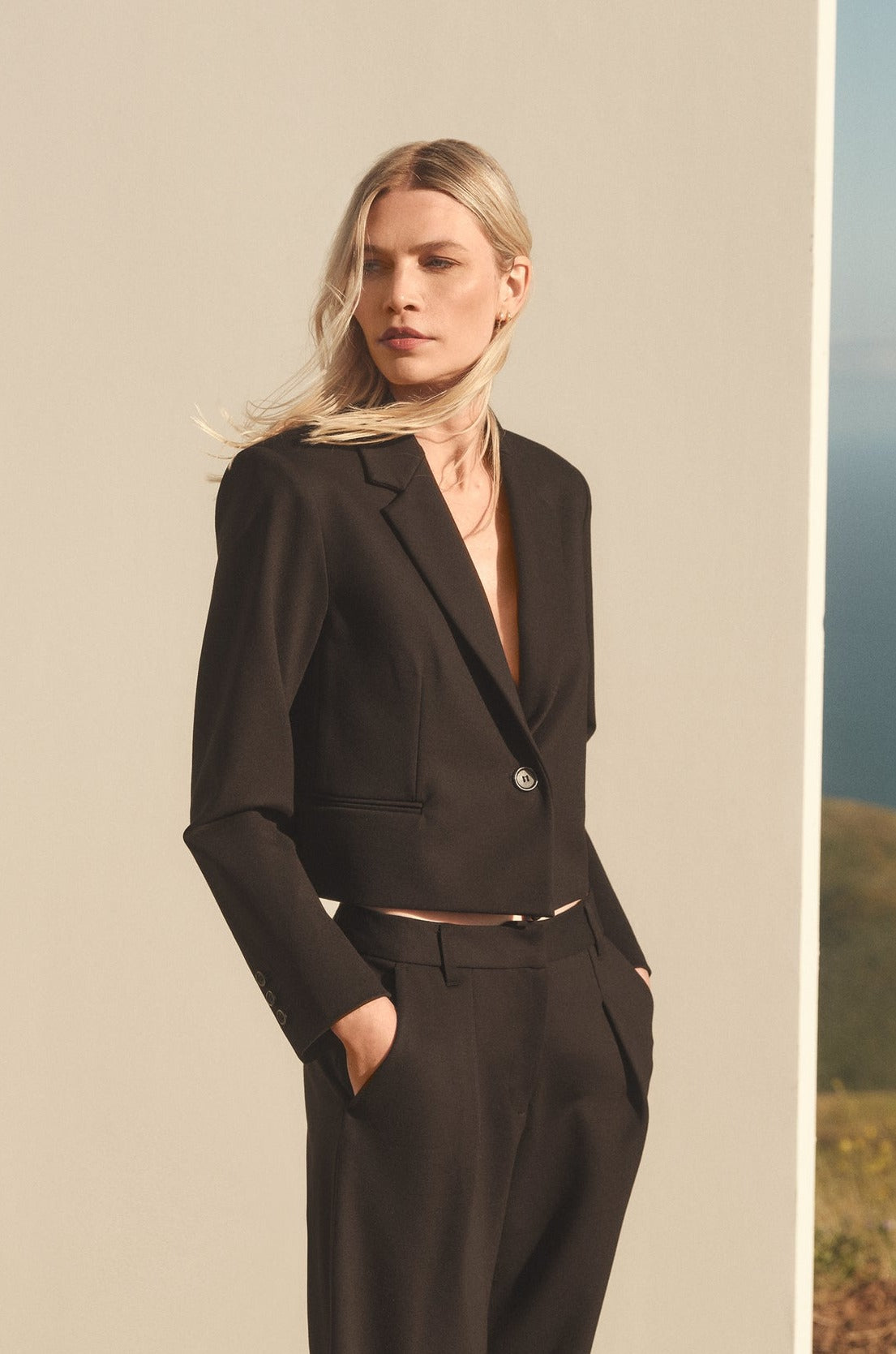 A person with long, blonde hair stands in front of a minimalist backdrop, wearing the contemporary ANYA PONTE CROPPED BLAZER by Velvet by Graham & Spencer and matching pants. With hands in pockets and a neutral expression, the single-button closure of the blazer adds an understated elegance to the outfit.-35655683670209