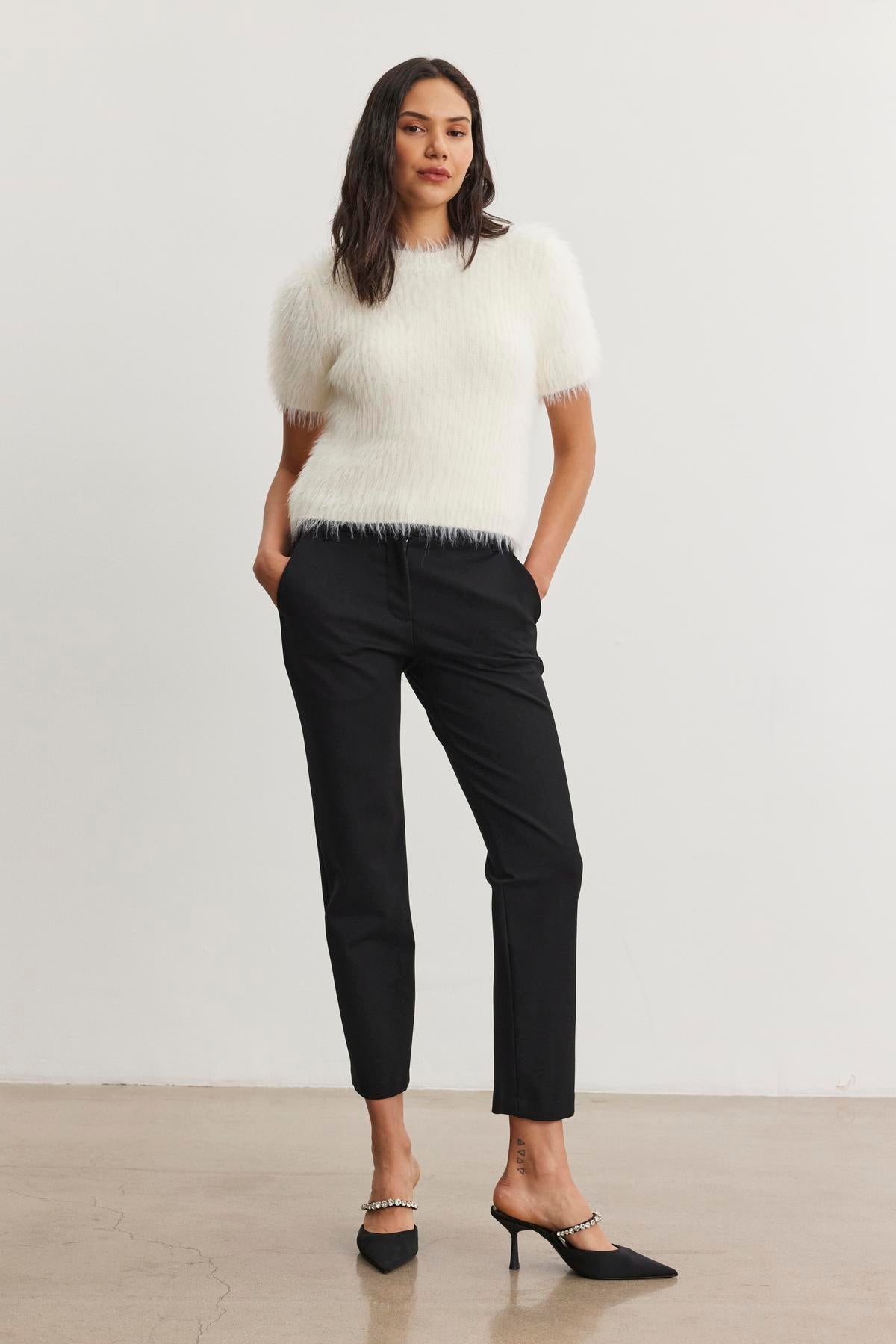 A person stands against a plain background, wearing a white fuzzy top paired with the JAY PONTE PANT in black from Velvet by Graham & Spencer, complemented by sleek black heels.-38246517702849