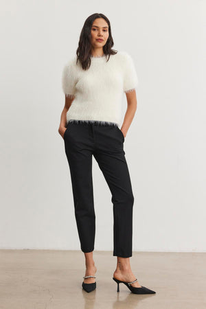 A person stands against a plain background, wearing a white fuzzy top paired with the JAY PONTE PANT in black from Velvet by Graham & Spencer, complemented by sleek black heels.