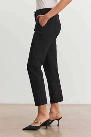 A person wearing a white top, the JAY PONTE PANT by Velvet by Graham & Spencer, and black high-heeled shoes featuring chain detail stands on a neutral background.