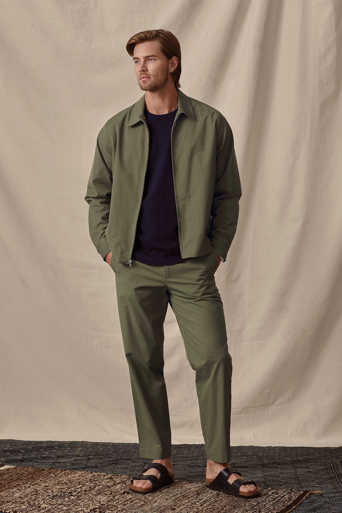 A man models a casual olive green Velvet by Graham & Spencer ALTON POPLIN ZIP-UP JACKET and trousers with a navy t-shirt and black sandals against a cream backdrop.-36299656298689