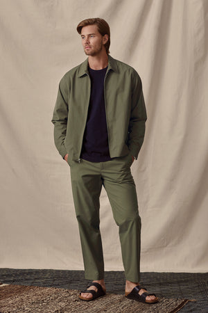 A man models a casual olive green Velvet by Graham & Spencer ALTON POPLIN ZIP-UP JACKET and trousers with a navy t-shirt and black sandals against a cream backdrop.