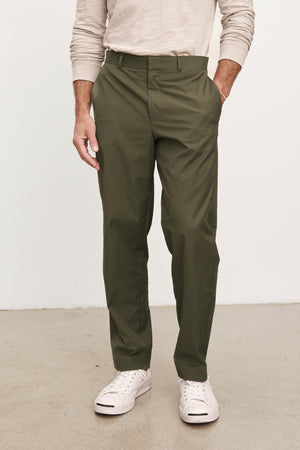A person wears Velvet by Graham & Spencer's STING POPLIN PANT in olive green cotton poplin, paired with a light beige long-sleeve shirt and white sneakers, standing on a light floor—an effortlessly versatile wardrobe staple.