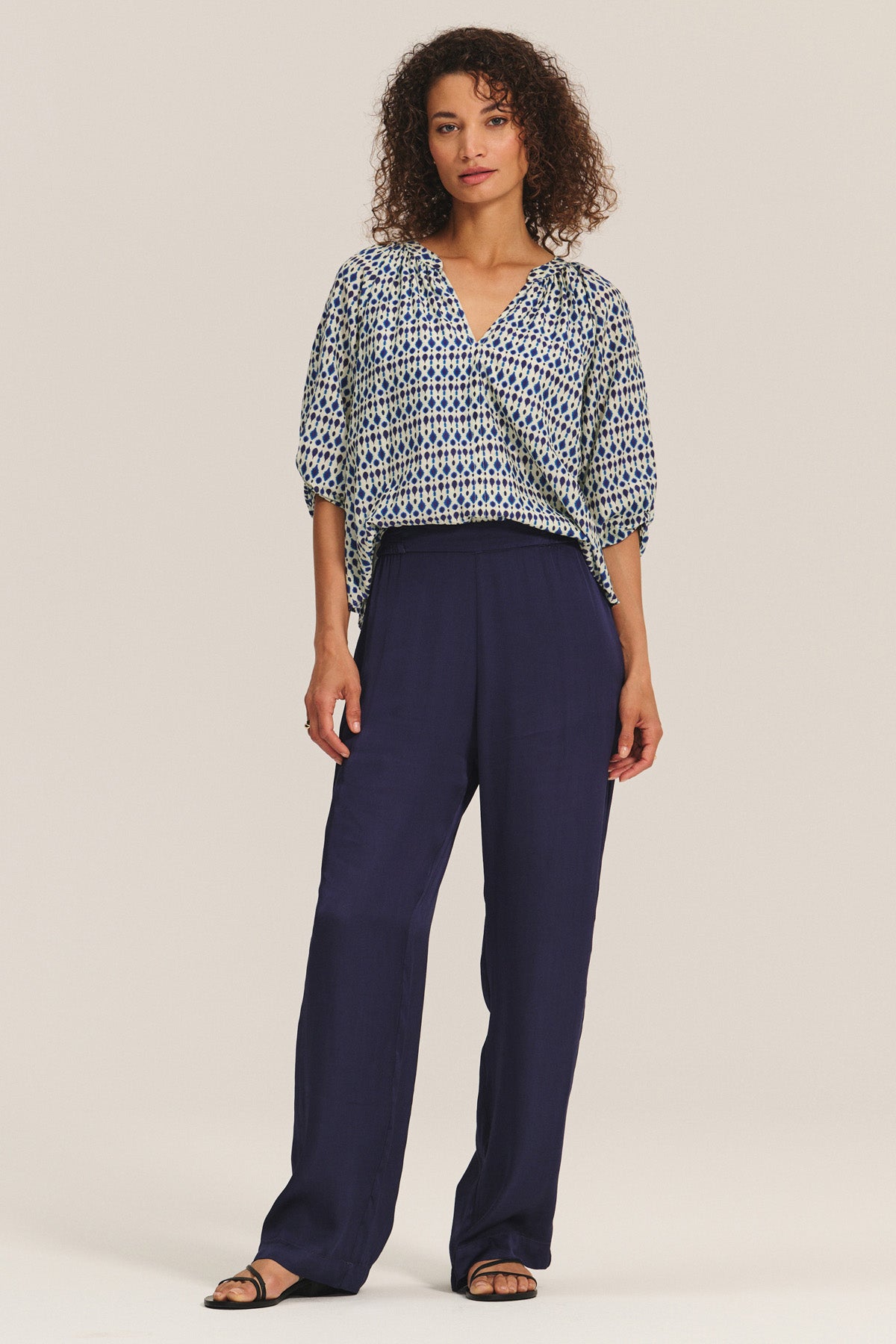 A woman wears the HARMONY BLOUSE by Velvet by Graham & Spencer—a flowy, printed cotton voile top with elbow-length sleeves—paired with loose navy trousers and black sandals, standing against a plain light background.-40732814966977
