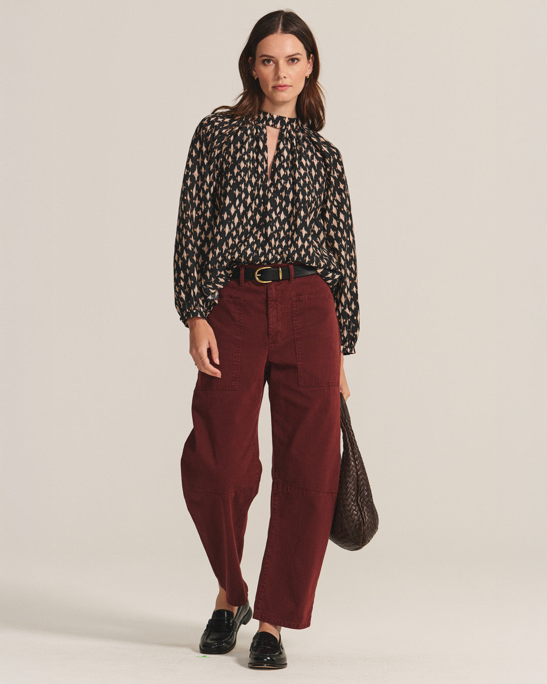 A woman stands facing forward wearing a black patterned blouse, black loafers, holding a textured brown bag, and maroon BRYLIE PANT by Velvet by Graham & Spencer featuring utilitarian patch pockets.-40247381590209