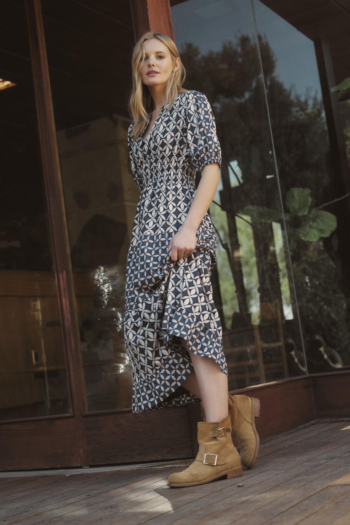 A woman in a JOEY DRESS by Velvet by Graham & Spencer, featuring a patterned, printed cotton cambric fabric and a v-neckline, stands in front of a glass door while holding the edge of her dress. She is wearing brown boots, and there are plants visible through the glass.