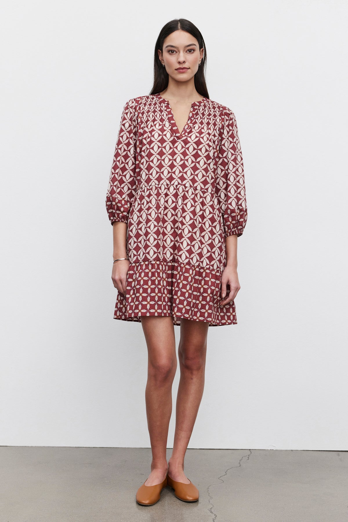 A woman stands against a plain background, showcasing the BECKIE DRESS from Velvet by Graham & Spencer. The lightweight cotton dress features a red and white geometric design with tiered detailing and smocked shoulder accents. It has three-quarter sleeves, and she accessorizes it with flat brown shoes. Her long dark hair completes the look.-37676900450497