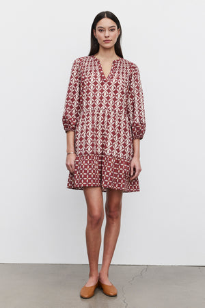 A woman stands against a plain background, showcasing the BECKIE DRESS from Velvet by Graham & Spencer. The lightweight cotton dress features a red and white geometric design with tiered detailing and smocked shoulder accents. It has three-quarter sleeves, and she accessorizes it with flat brown shoes. Her long dark hair completes the look.