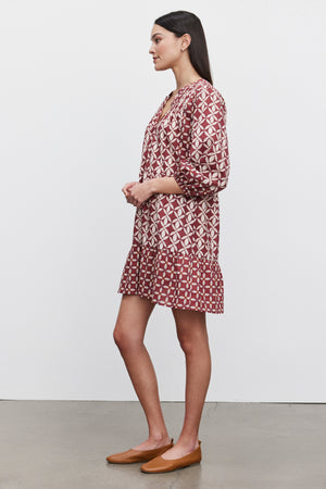 An individual wearing the BECKIE DRESS by Velvet by Graham & Spencer, a patterned, knee-length, lightweight cotton dress featuring smocked shoulder detailing and a ruffled hem, paired with tan flats, stands sideways against a plain background.