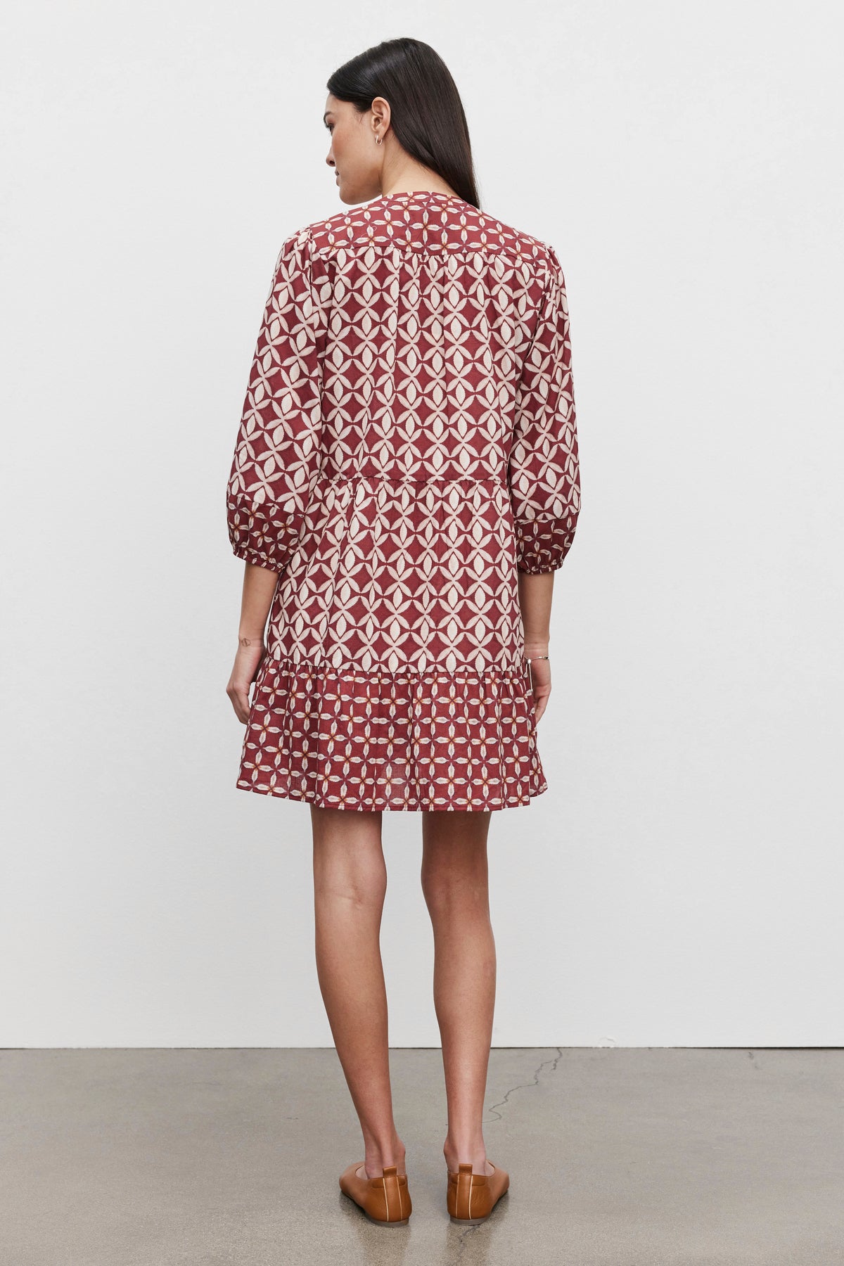 A woman stands facing away, wearing the BECKIE DRESS by Velvet by Graham & Spencer. The dress features a patterned red, tiered design with three-quarter sleeves and is paired with brown shoes. She has long dark hair and is posed against a plain white background on a cement floor.-37676900384961