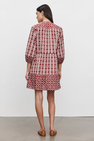 A woman stands facing away, wearing the BECKIE DRESS by Velvet by Graham & Spencer. The dress features a patterned red, tiered design with three-quarter sleeves and is paired with brown shoes. She has long dark hair and is posed against a plain white background on a cement floor.