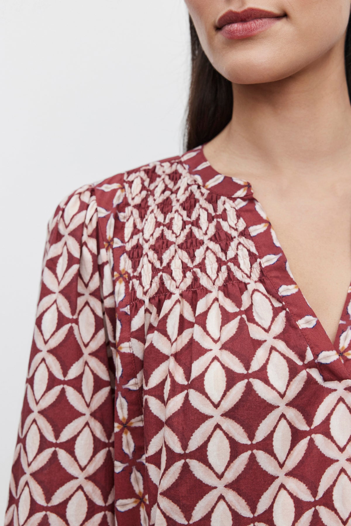 Close-up of a person wearing the BECKIE DRESS by Velvet by Graham & Spencer, a maroon blouse with a white geometric pattern and pleated detailing around the neckline, reminiscent of the intricate designs found in tiered design dresses, shown from mid-chest to just above the lips.