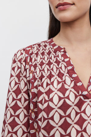 Close-up of a person wearing the BECKIE DRESS by Velvet by Graham & Spencer, a maroon blouse with a white geometric pattern and pleated detailing around the neckline, reminiscent of the intricate designs found in tiered design dresses, shown from mid-chest to just above the lips.