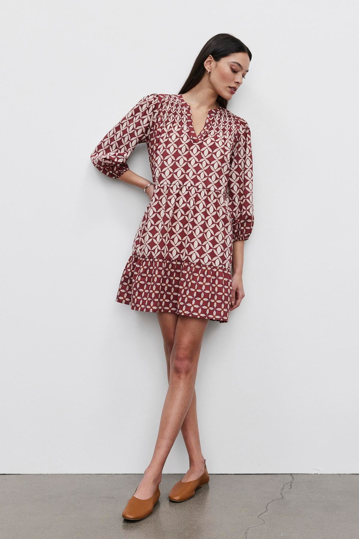 A woman wearing the BECKIE DRESS from Velvet by Graham & Spencer, featuring a maroon, tiered design with three-quarter sleeves, stands against a plain white wall. She is also wearing tan loafers and has her arms slightly crossed.